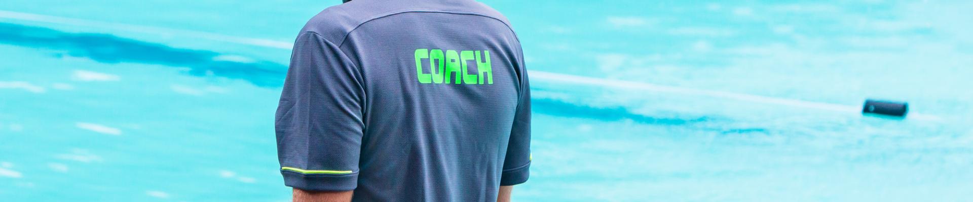 R&uuml;ckansicht eines Trainers mit der Aufschrift "COACH" auf dem Shirt vor einem Schwimmbecken.