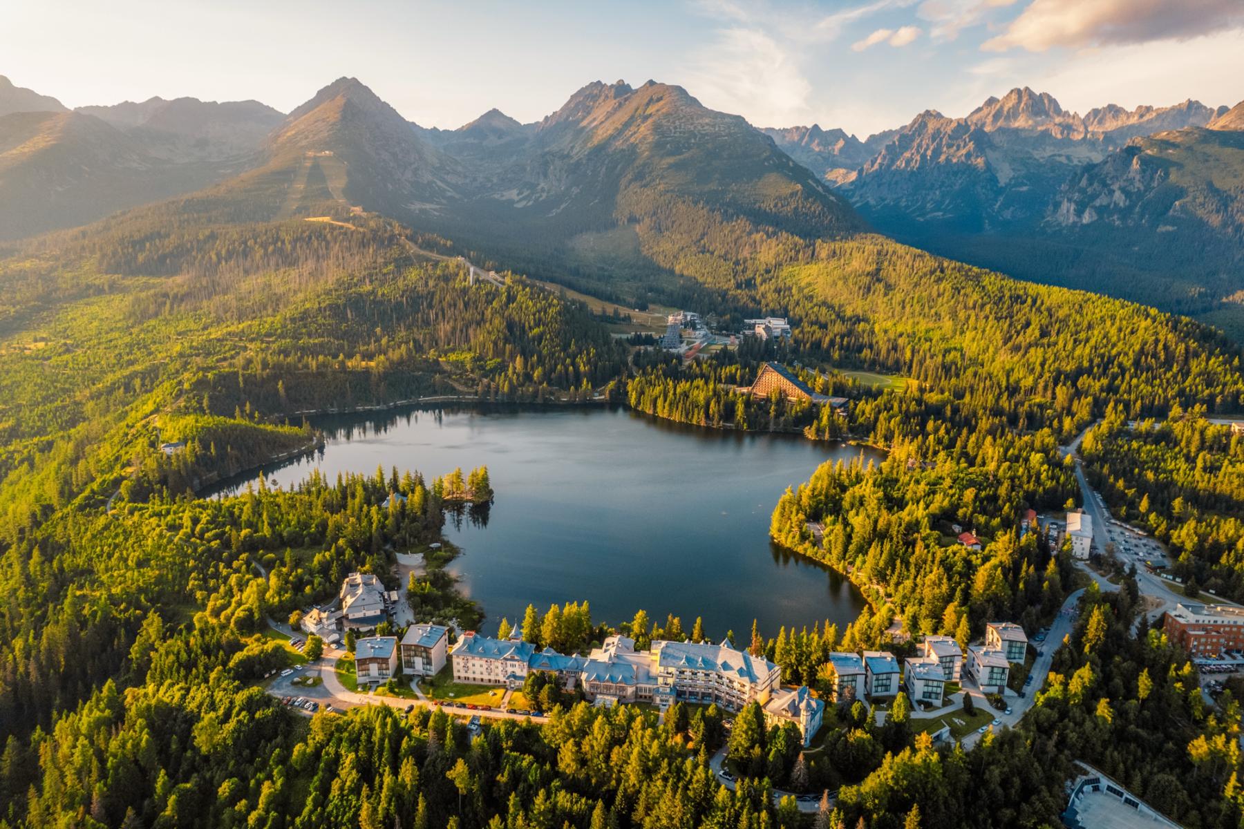 Malerischer Bergsee in der Slowakei, umgeben von dichten W&auml;ldern und imposanten Bergen, mit Hotels und Geb&auml;uden am Ufer in warmem Licht