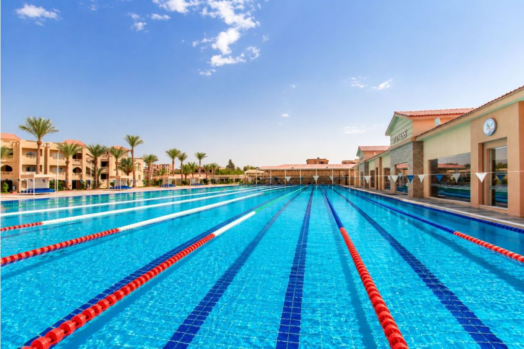 Ein olympisches Freibad mit klarem, blauem Wasser und Schwimmbahnmarkierungen, umgeben von Palmen, sonnendurchfluteten Gebäuden und einem strahlend blauen Himmel mit wenigen Wolken.