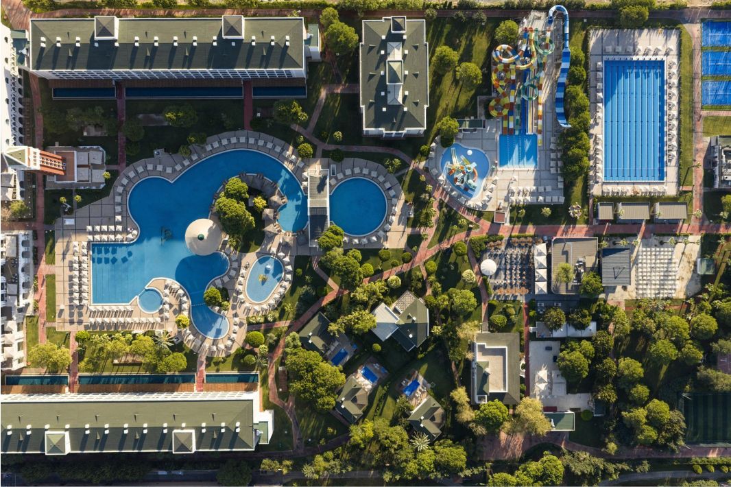 Luftaufnahme eines Resorts mit großen, einzigartig geformten Swimmingpools, einer hohen Wasserrutschenstruktur, einem rechteckigen Pool, Sonnenliegen, grünen Rasenflächen, Gehwegen und umliegenden Gebäuden mit Bäumen auf dem Gelände.