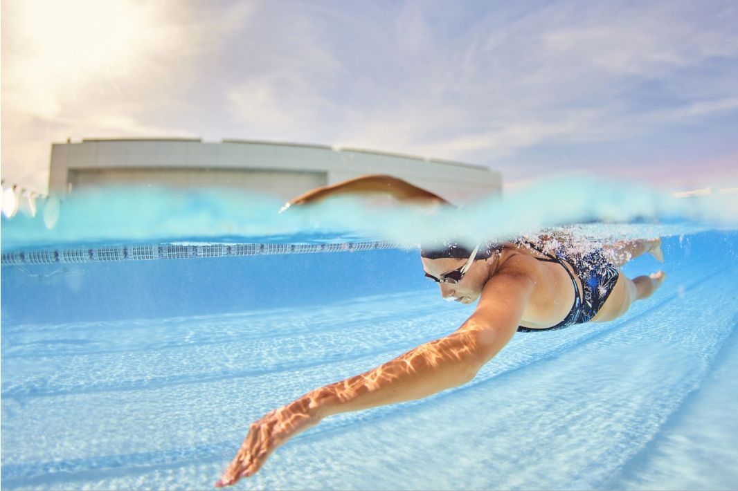 Ein Schwimmer in Badeanzug, Schwimmbrille und Badekappe führt in einem klaren Freibad den Freistilschwimmzug aus, der teilweise unter Wasser mit Sonnenlicht und einem Gebäude im Hintergrund aufgenommen wurde.