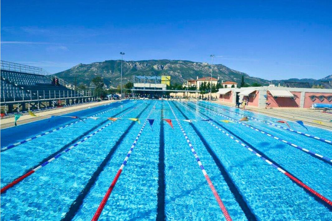 Ein olympisches Außenschwimmbecken mit Bahntrennern, umgeben von Tribünen und Gebäuden, vor der Kulisse der Berge und unter einem strahlend blauen Himmel.