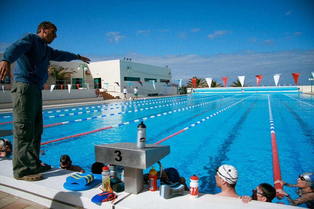 Ein Trainer gibt Anweisungen an eine Gruppe von Schwimmern am Rand eines olympischen Außenpools mit roten und blauen Bahnmarkierungen unter blauem Himmel.