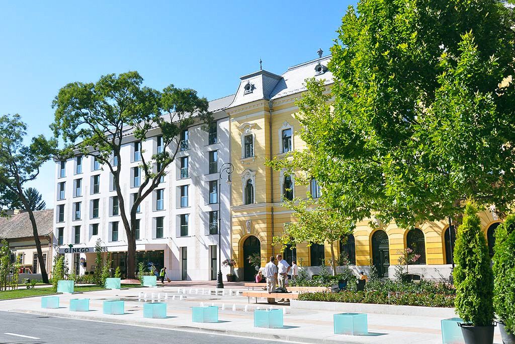 Außenansicht des Hotels Gingko mit einer modernen Fassade und einem historischen Flügel, umgeben von grünen Bäumen und einem gepflegten Platz.