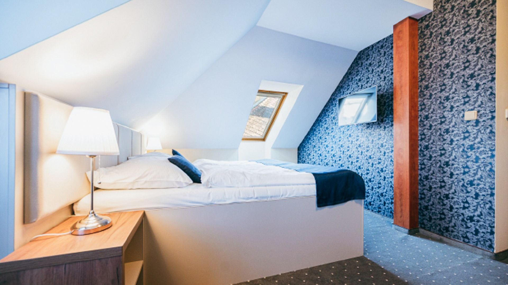 Gemütliches Hotelzimmer mit einem Doppelbett, Dachschräge und minimalistischer Einrichtung in Blau- und Weißtönen.
