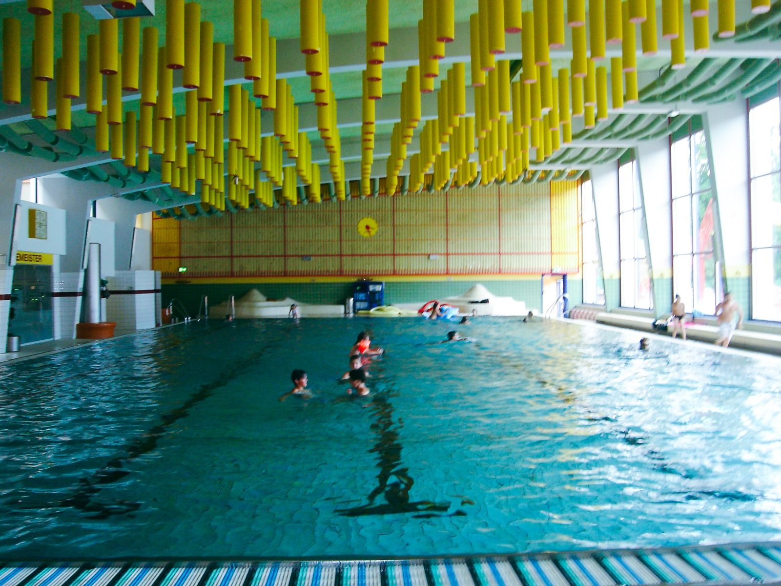 Große Innenansicht des Schwimmbereichs mit auffälliger gelber Deckengestaltung und Menschen im Wasser.