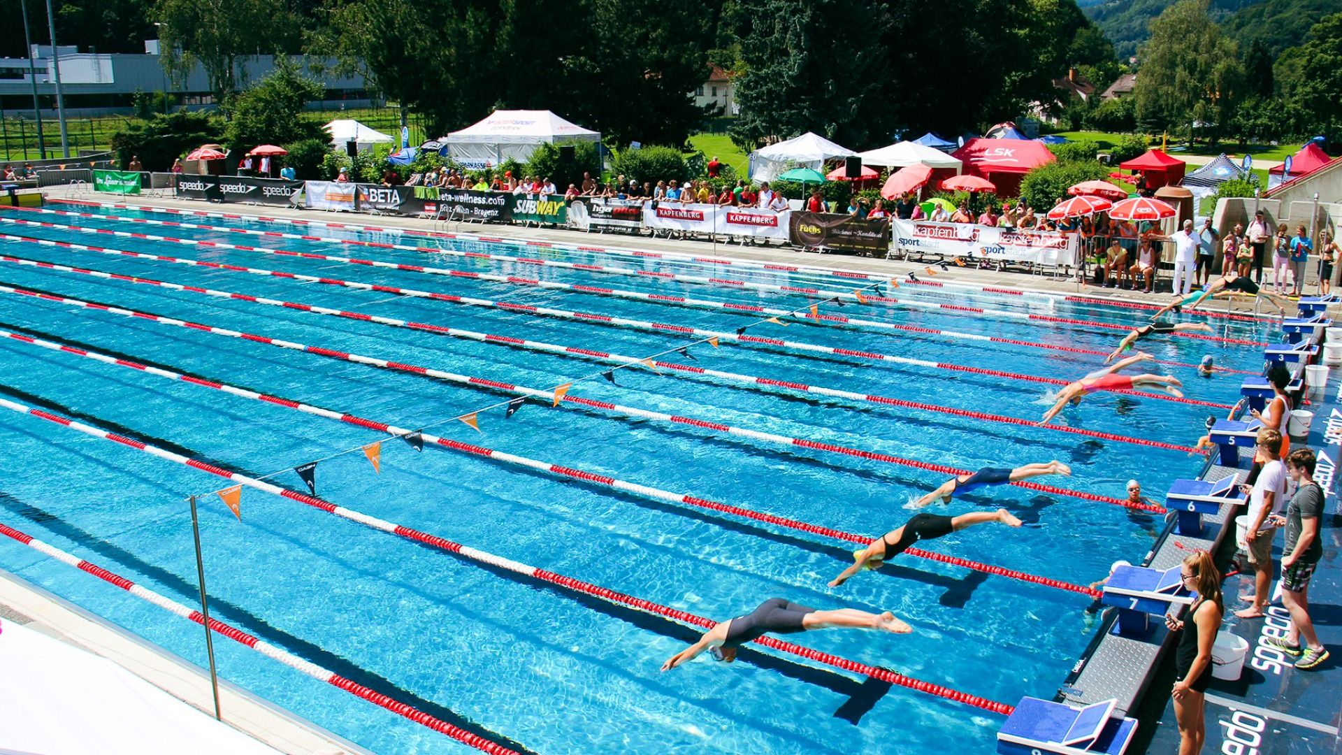 Schwimmwettkampf im Freibad des Sporthotel Kapfenberg, mehrere Schwimmer starten ins Wasser, während Zuschauer unter Sonnenschirmen zuschauen