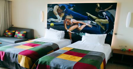 Hotelgeb&auml;ude mit mehreren Balkonen, neben einer roten Laufbahn und umgeben von Palmen.