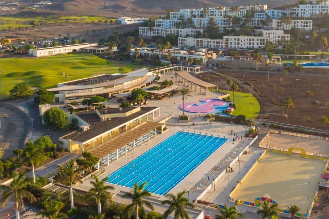 Luftaufnahme eines modernen Sportkomplexes mit einem großen Freibad, angrenzenden Palmen, angelegten Rasenflächen, einem Sand-Sportplatz, einem bunten Spielplatz und umliegenden Wohngebäuden.