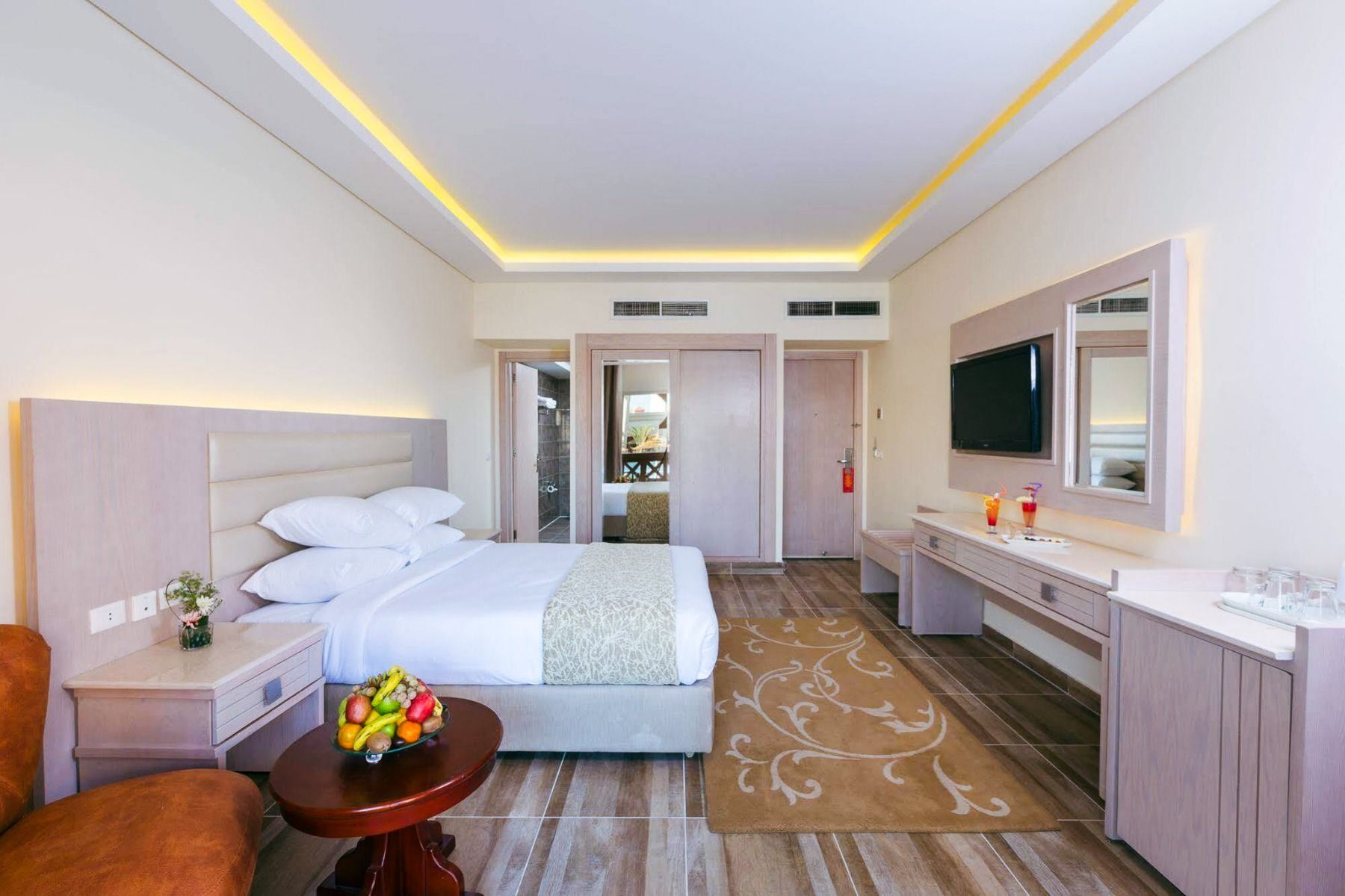 Aqua Vista Resort offers a modern hotel room with a neatly made double bed, cushioned headboard, warm lighting, wooden side table with fruit bowl, large TV, and desk with mirror—ideal for ein erfolgreiches Trainingslager in den Wintermonaten.
