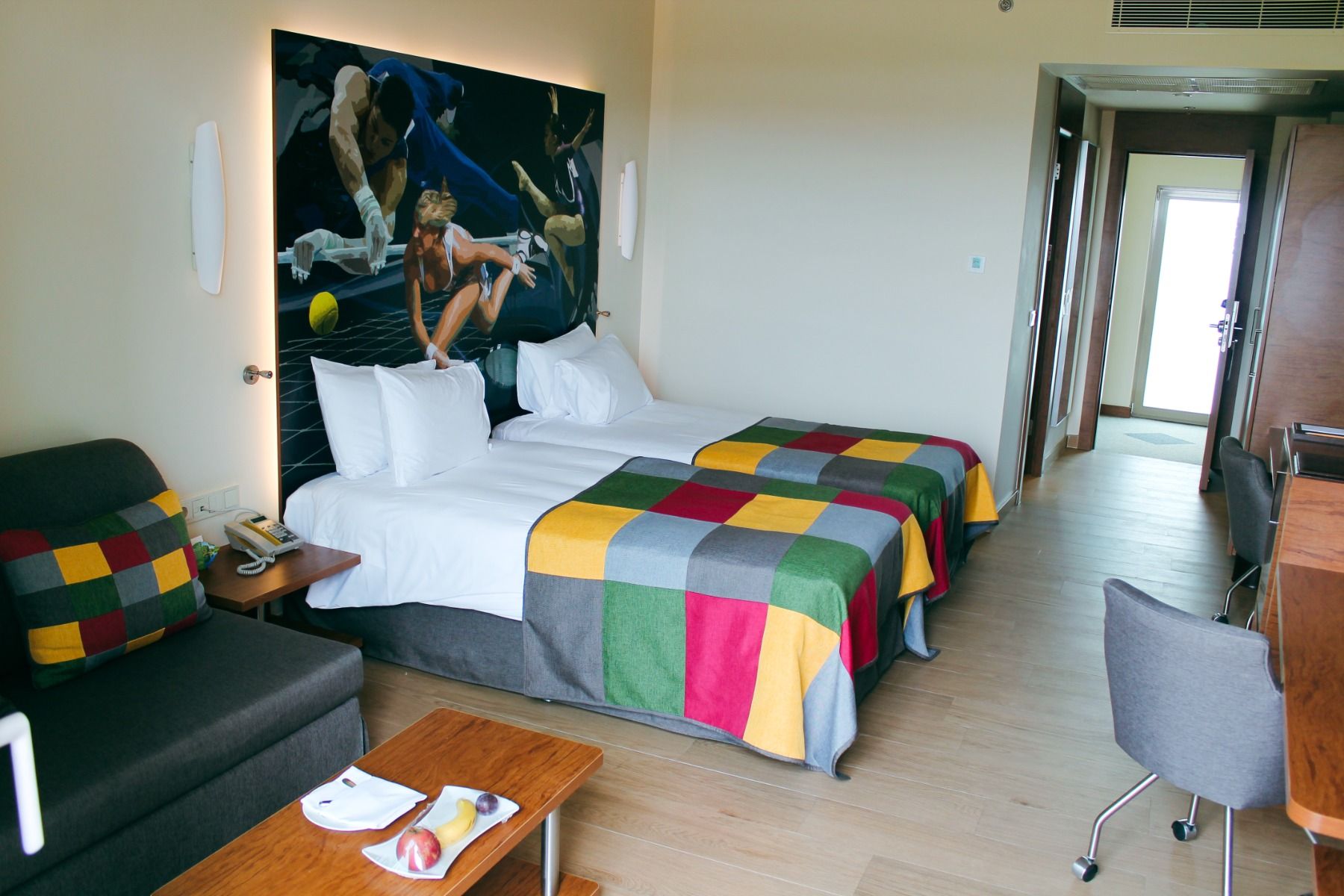 Modernes Hotelzimmer mit zwei Einzelbetten, farbenfroher Tagesdecke und einer Sport-Collage an der Wand.
