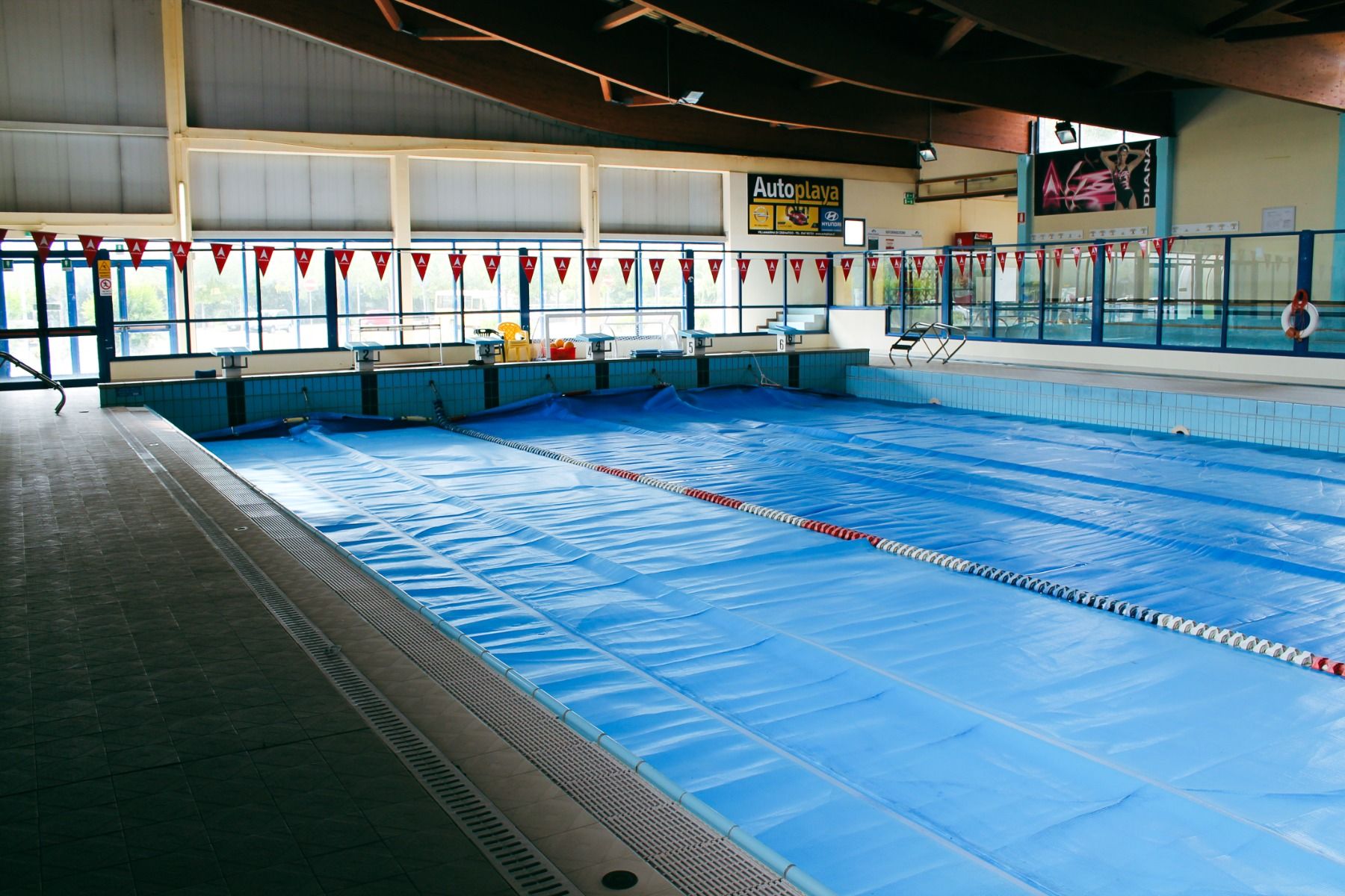 Hallenbad mit blau abgedeckten Schwimmbahnen, großen Fenstern, roten Wimpeln und seitlichen Startblöcken