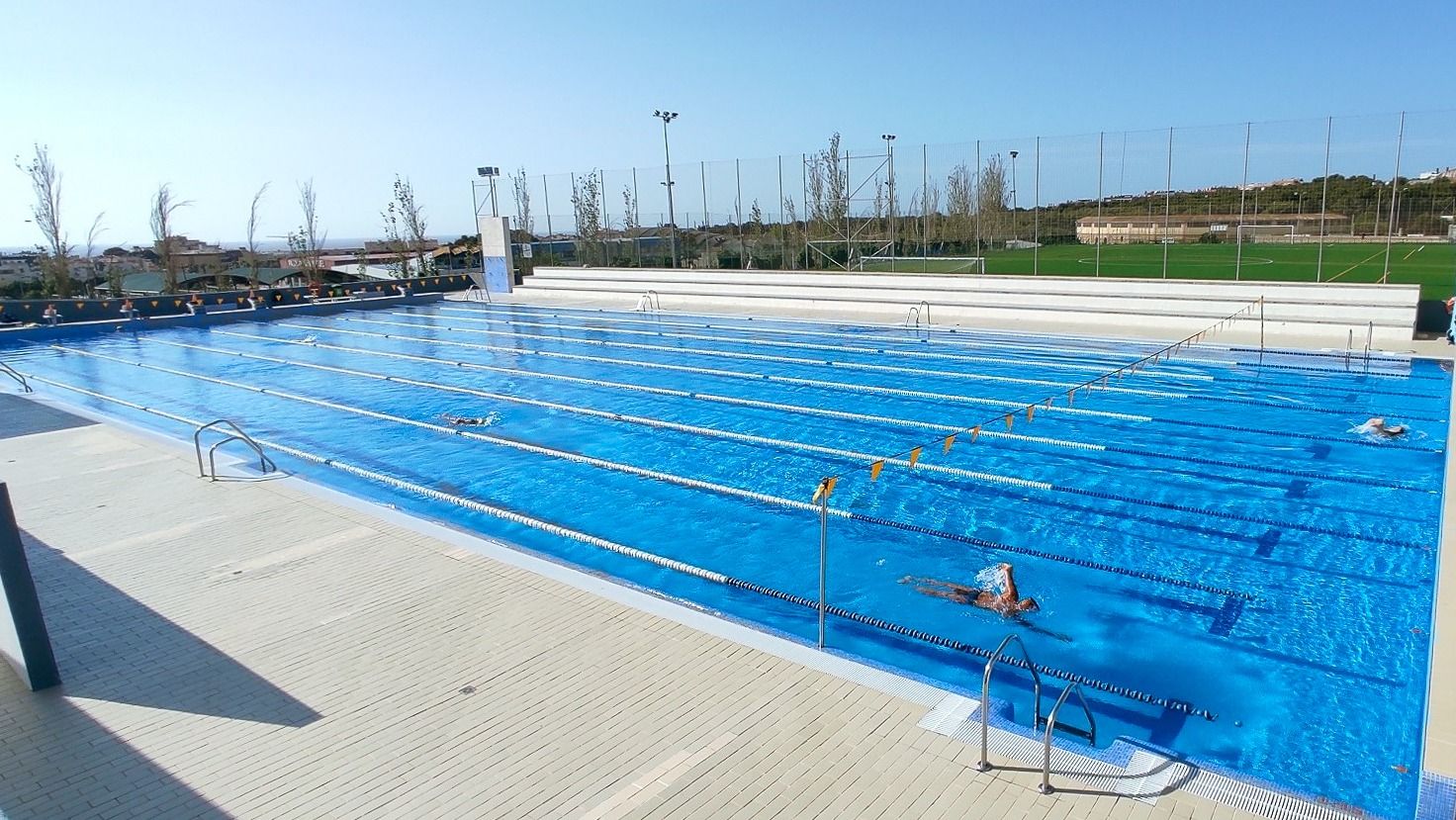 Olympisches Freiluft-Schwimmbecken mit mehreren Bahnen, Schwimmer im Wasser und Sportplatz im Hintergrund.