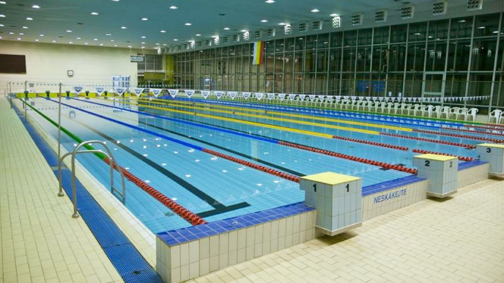 Blick auf das helle, geräumige Schwimmbecken mit klar abgetrennten Bahnen, großen Fenstern und Sitzbereichen an der Seite.