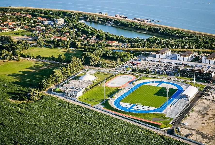 Luftaufnahme des Leichtathletikstadions mit blauer Laufbahn, Fußballfeld und Sportanlagen am Flussufer.