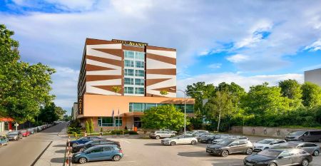 Ein modernes Hotelgeb&auml;ude mit auff&auml;lligen geometrischen Mustern in Braun und Wei&szlig;, umgeben von gr&uuml;nen B&auml;umen und einem gro&szlig;en Parkplatz im Vordergrund.