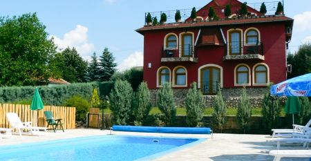 Rotes Hotelgeb&auml;ude mit dekorativen Balkonen, einem Pool und gepflegter Gartenlandschaft.