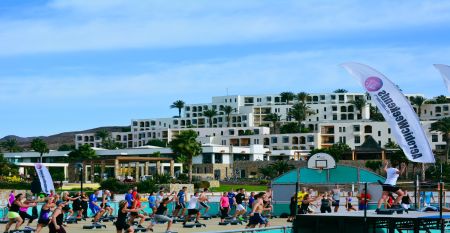 Outdoor-Fitnesskurs mit einer Gruppe von Menschen, die gemeinsam auf Steppern trainieren, vor der Kulisse einer wei&szlig;en, terrassenf&ouml;rmigen Hotelanlage.