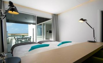Moderne weiße Hotelvilla mit Panoramafenstern und überdachter Terrasse, umgeben von schwarzem Sand und Blick auf eine ruhige Lagune.
