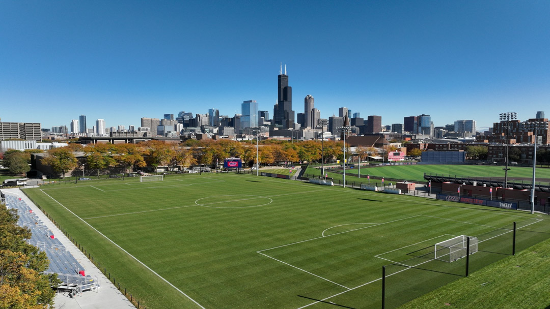 Fußballfeld mit gepflegtem Rasen vor einer modernen Skyline, umgeben von Flutlichtmasten und Tribünen unter klarem, blauem Himmel.