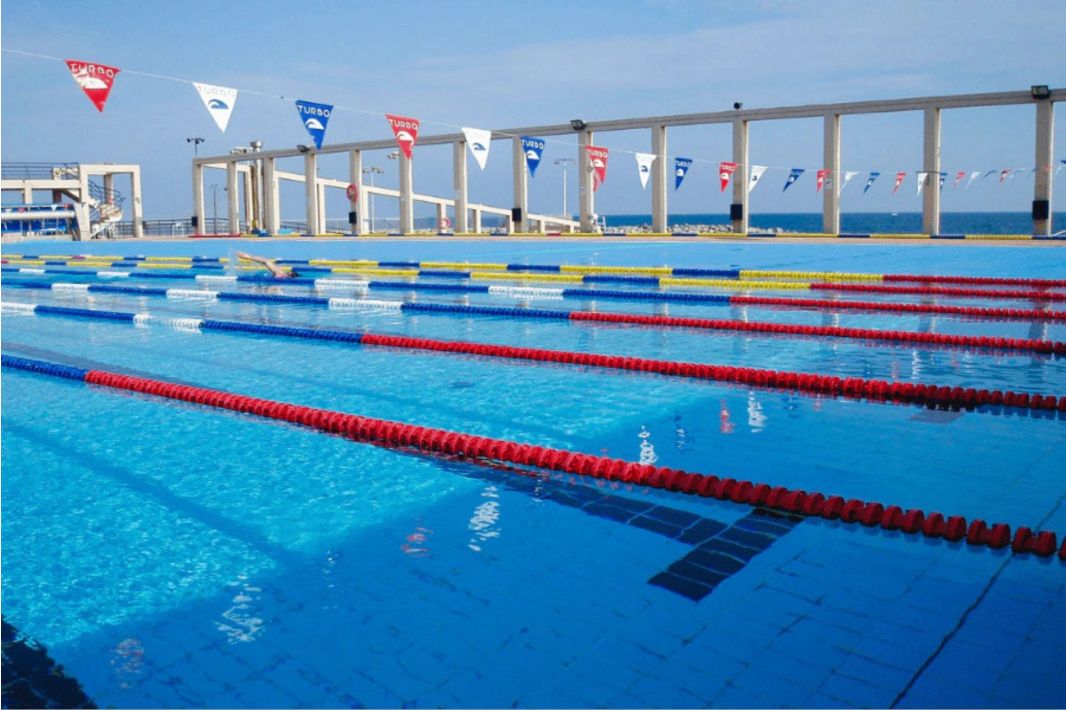 Freibad mit mehreren Bahnen, die durch rote, blaue und gelbe Markierungen unterteilt sind, weißen Säulen am Beckenrand und dreieckigen Fahnen, die über dem Becken hängen. Im Hintergrund sind das Meer und der Himmel zu sehen.