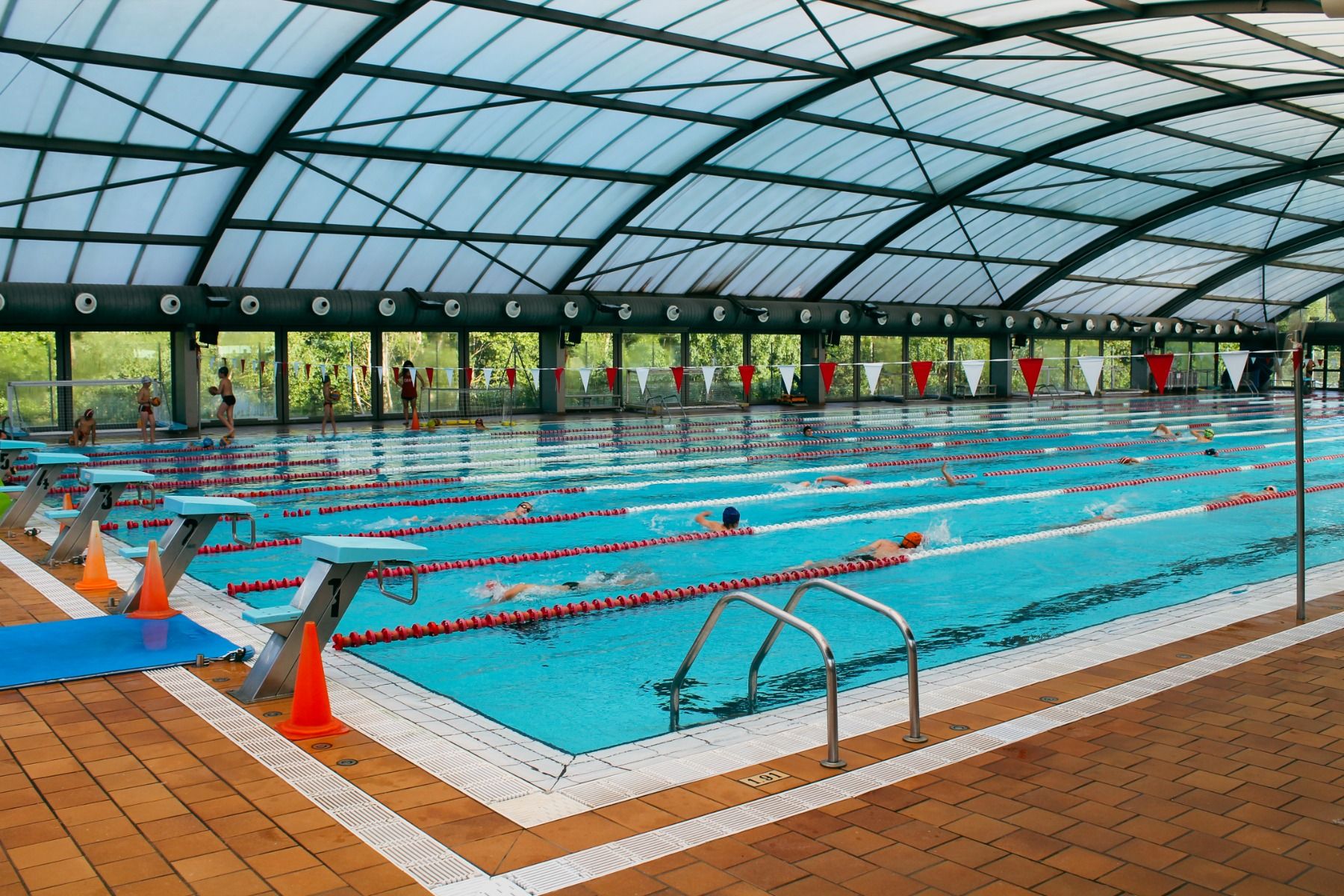 Ein großes, überdachtes Schwimmbecken mit mehreren Bahnen, in denen Schwimmer trainieren. Die Poolumrandung ist mit orangen Pylonen und Startblöcken ausgestattet.