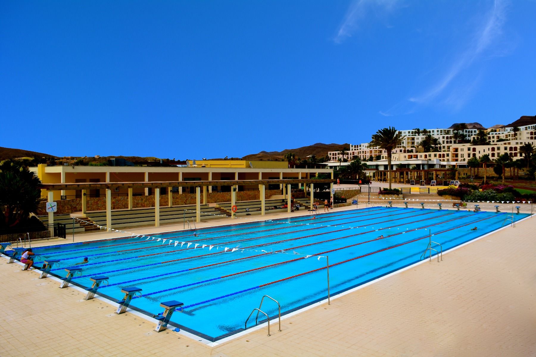 Ein weitläufiges Außen-Schwimmbecken mit mehreren Bahnen und blauen Startblöcken, umgeben von Sitzplätzen und einer modernen Hotelanlage im Hintergrund.