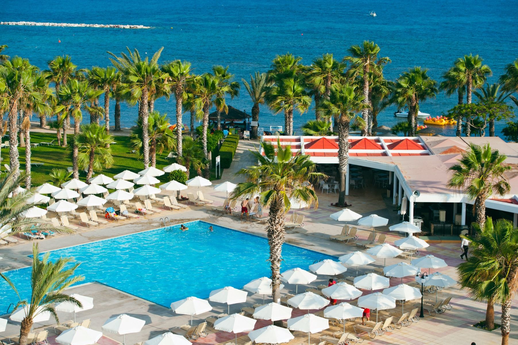 Blick auf das Außenpool-Areal und den angrenzenden Strand mit Liegen und Sonnenschirmen am Louis Phaethon Beach Hotel.
