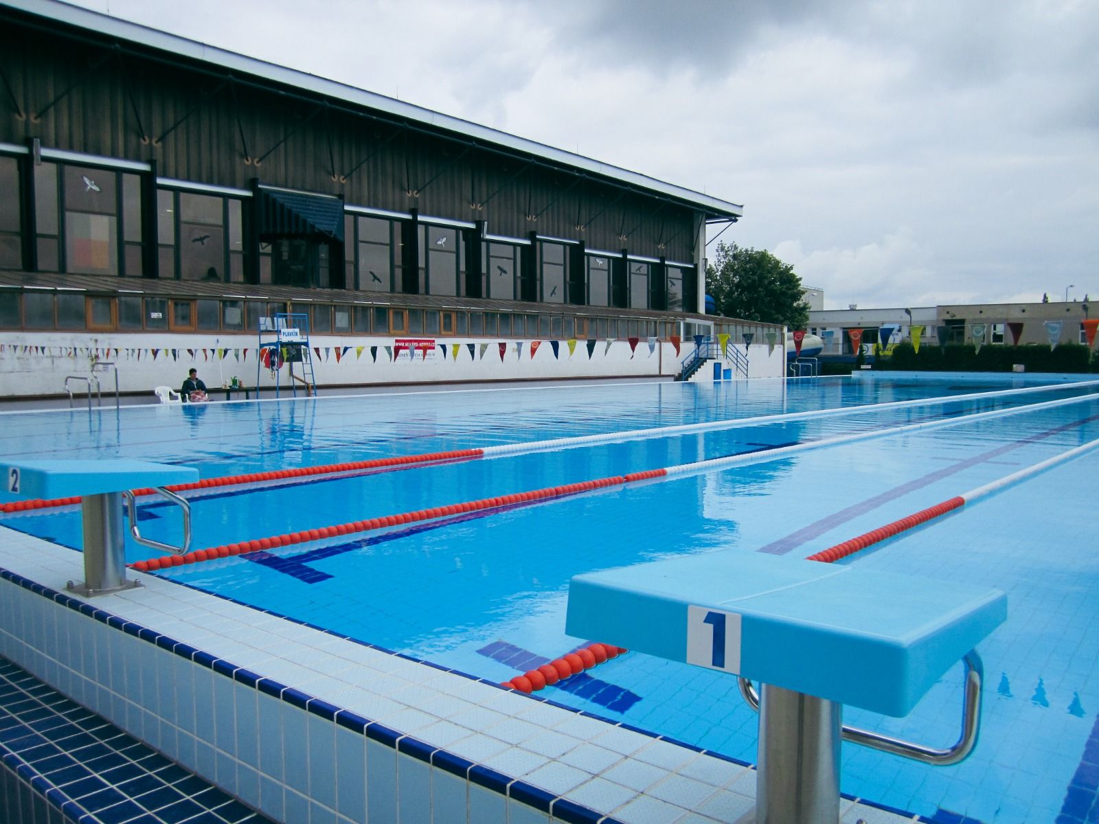 Außenaufnahme eines Wettkampfschwimmbads mit Startblöcken und einem angrenzenden Gebäude bei bewölktem Himmel.