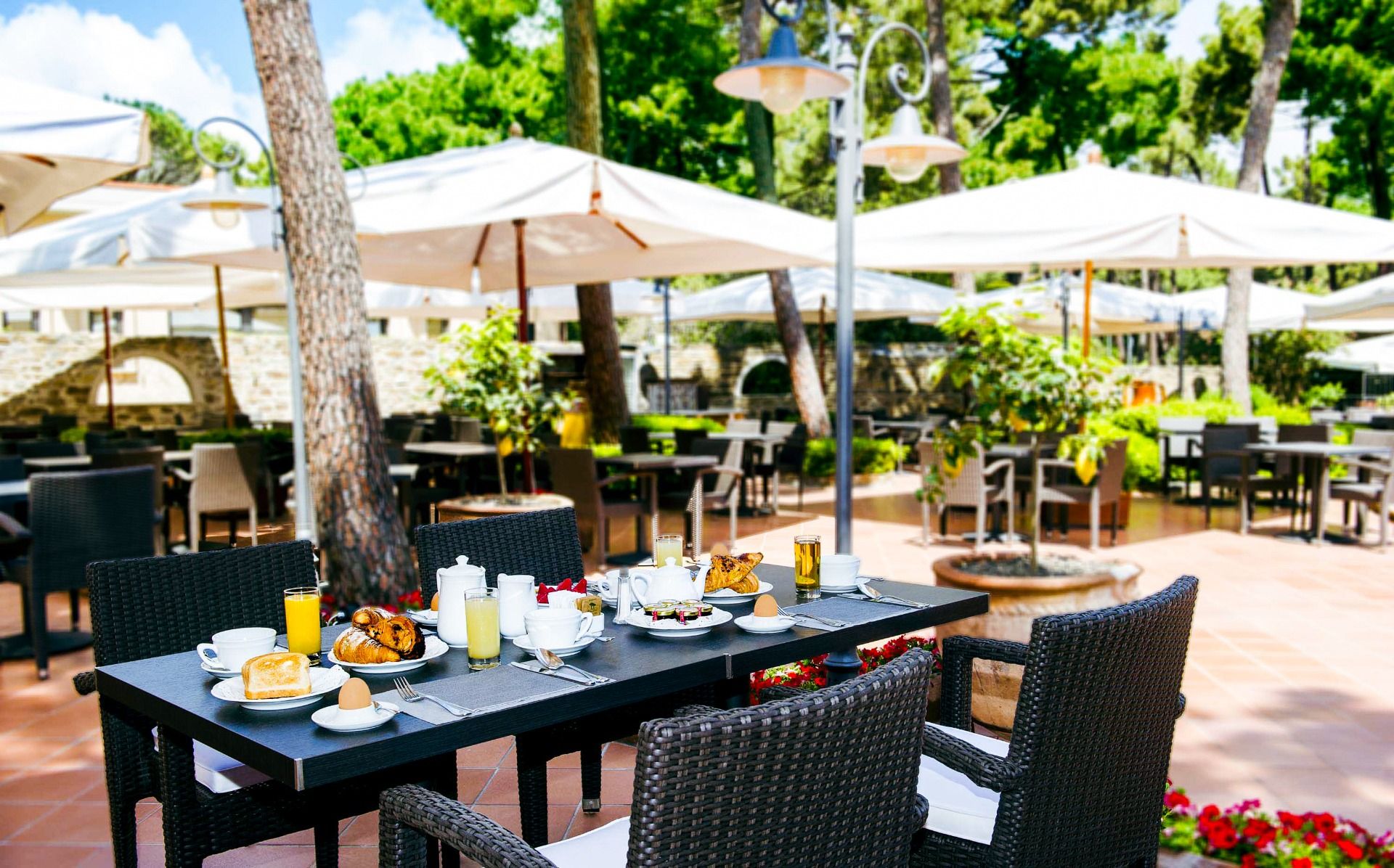 Außenbereich des Restaurants des Hotels Riva del Sole mit Rattanmöbeln unter großen Sonnenschirmen und Pinienbäumen
