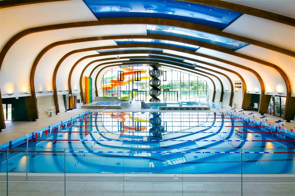 Großes, lichtdurchflutetes Hallenbad mit Wettkampf-Schwimmbecken, Wasserrutschen und gebogenem Holzdach.