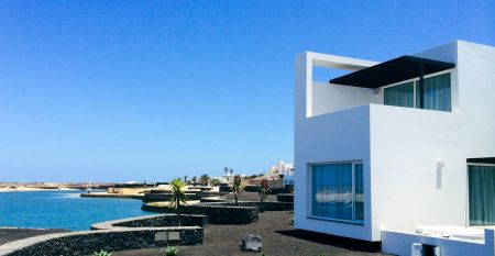Moderne wei&szlig;e Hotelvilla mit Panoramafenstern und &uuml;berdachter Terrasse, umgeben von schwarzem Sand und Blick auf eine ruhige Lagune.