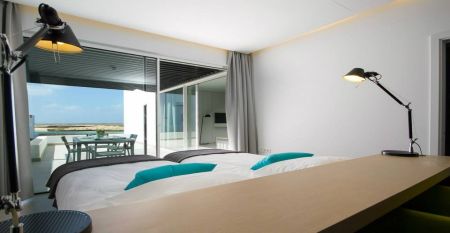 Moderne wei&szlig;e Hotelvilla mit Panoramafenstern und &uuml;berdachter Terrasse, umgeben von schwarzem Sand und Blick auf eine ruhige Lagune.