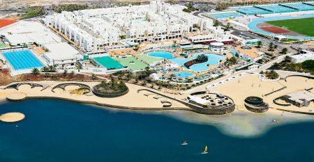 Moderne wei&szlig;e Hotelvilla mit Panoramafenstern und &uuml;berdachter Terrasse, umgeben von schwarzem Sand und Blick auf eine ruhige Lagune.