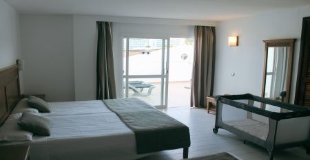Hotelgebäude im mediterranen Stil mit weißen Balkonen, Palmen und einem großen Poolbereich mit Sonnenliegen und Sonnenschirmen.