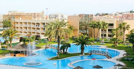 Weitl&auml;ufiger Hotelgarten mit geschwungenem Pool, Palmen und einer Br&uuml;cke, umgeben von Hotelgeb&auml;uden.