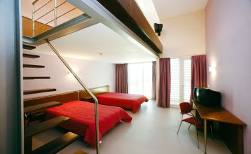 Helles Hotelzimmer mit zwei Betten mit einer roten Tagesdecke.  Zudem eine Treppe, die zu einer zweiten Ebene im Zimmer führt.