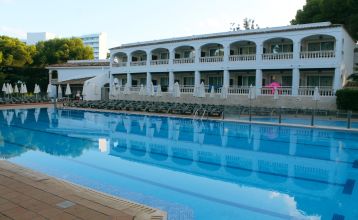 Hotelgebäude im mediterranen Stil mit weißen Balkonen, Palmen und einem großen Poolbereich mit Sonnenliegen und Sonnenschirmen.