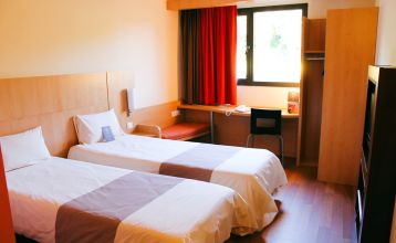 Außenansicht des Ibis Hotels mit moderner, grauer Fassade und blauer „Ibis Budget“- sowie roter „Ibis Hotel“-Beschriftung vor einem klaren blauen Himmel.