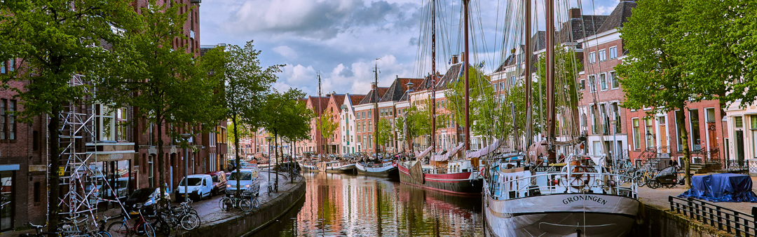 In Groningen, Niederlande, liegen Boote unter einem teilweise bewölkten Himmel an einem Kanal, der von Bäumen und historischen Backsteingebäuden gesäumt ist.